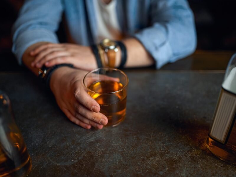A man's hands grip a glass of whiskey on a dark bar counter with bottles nearby.