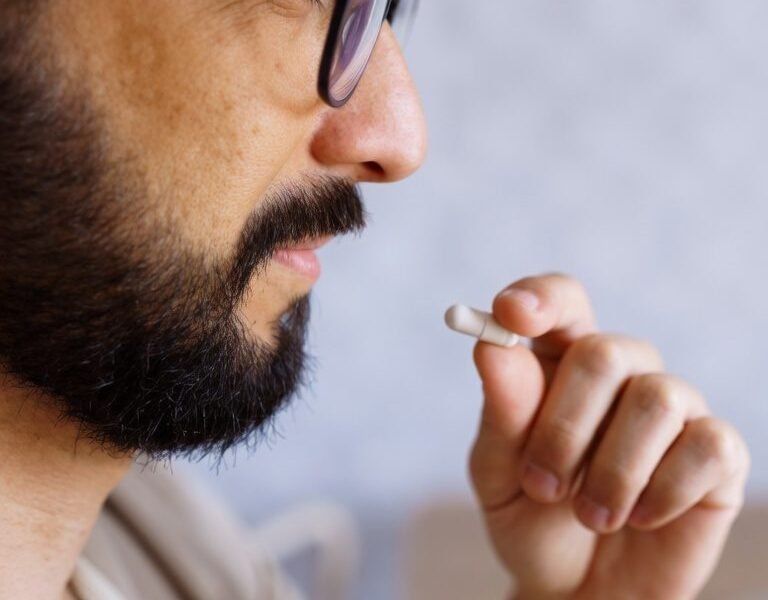 A man with glasses holds a small white capsule up toward his mouth, about to take it.