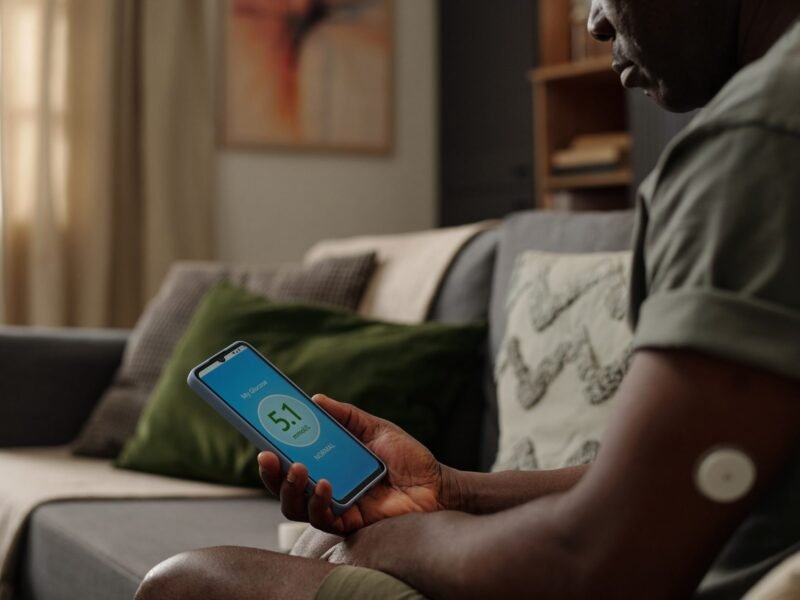 Man sitting on a couch checking a CGM glucose reading on his smartphone while wearing an arm sensor.