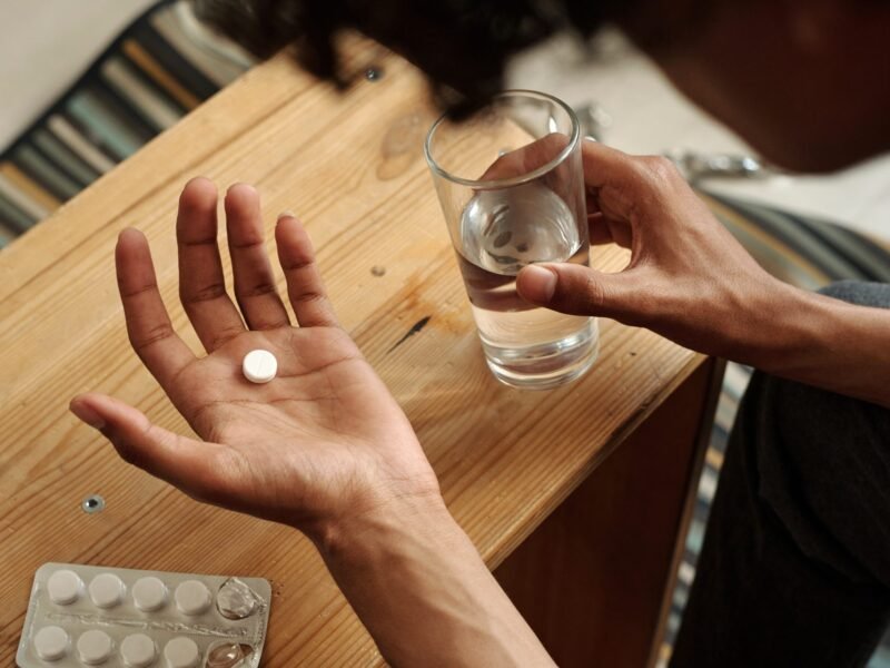Person holding a single white pill in an open palm alongside a glass of water at a wooden table.