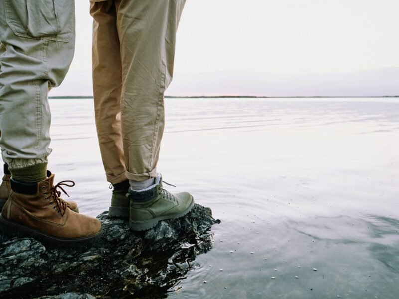 Two people standing on rocks at the edge of a calm body of water, shown from the knees down.