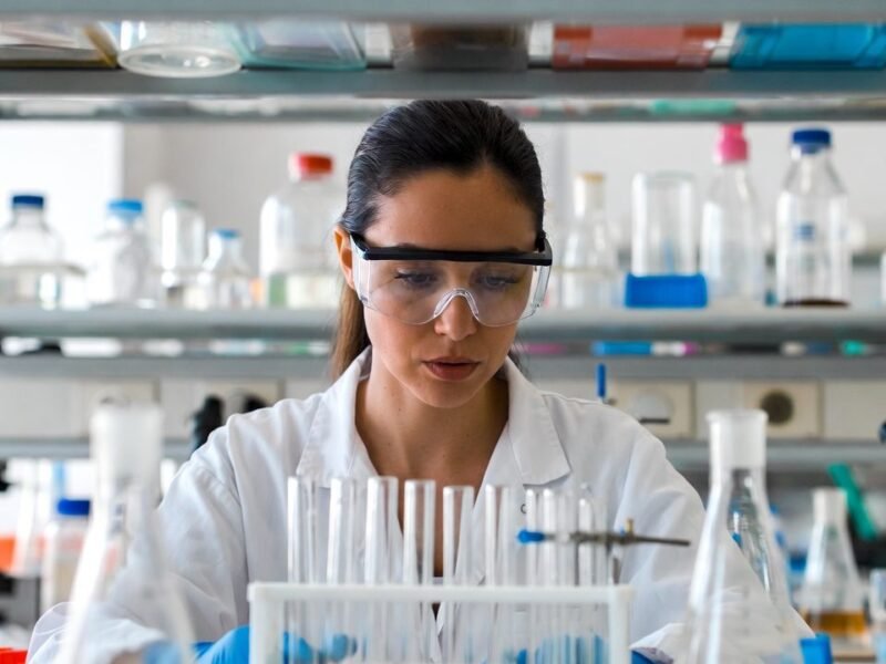 Female scientist in safety goggles examining test tubes inside a laboratory.
