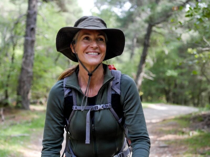 Middle-aged woman smiling while hiking on a wooded trail with a large backpack.