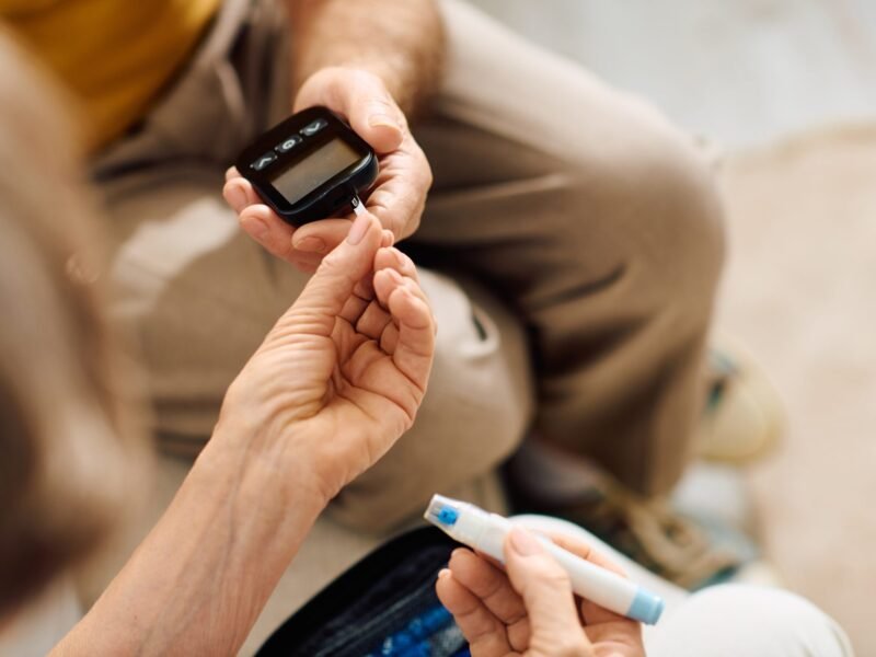 Close-up of a person assisting a senior with a blood glucose meter and lancet device.