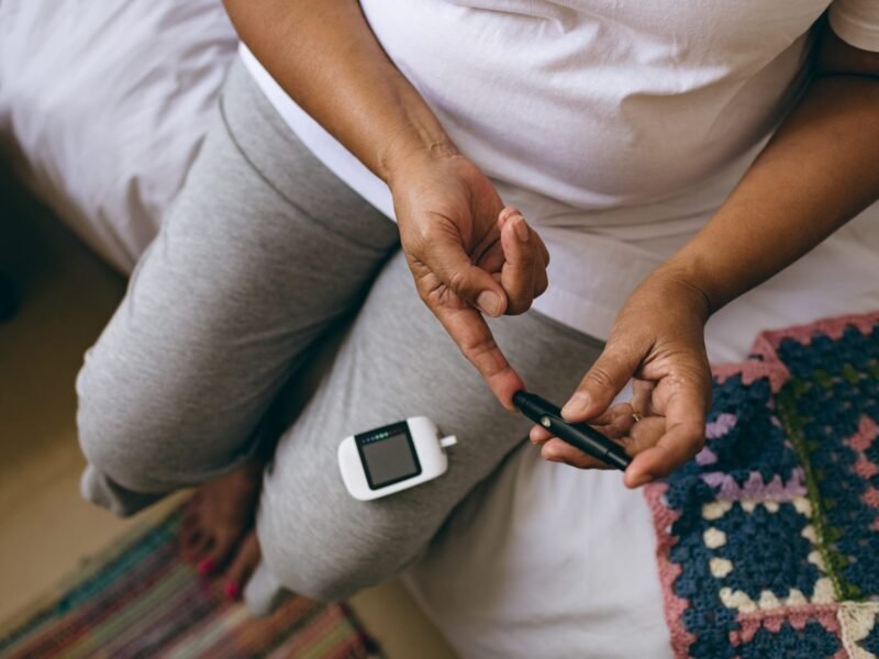 Woman sitting on a bed using a lancet to check her blood sugar with a glucose meter on her lap.