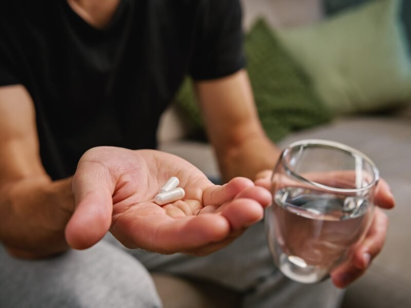 Man's open palm holding two white capsules beside a glass of water, seated on a couch.