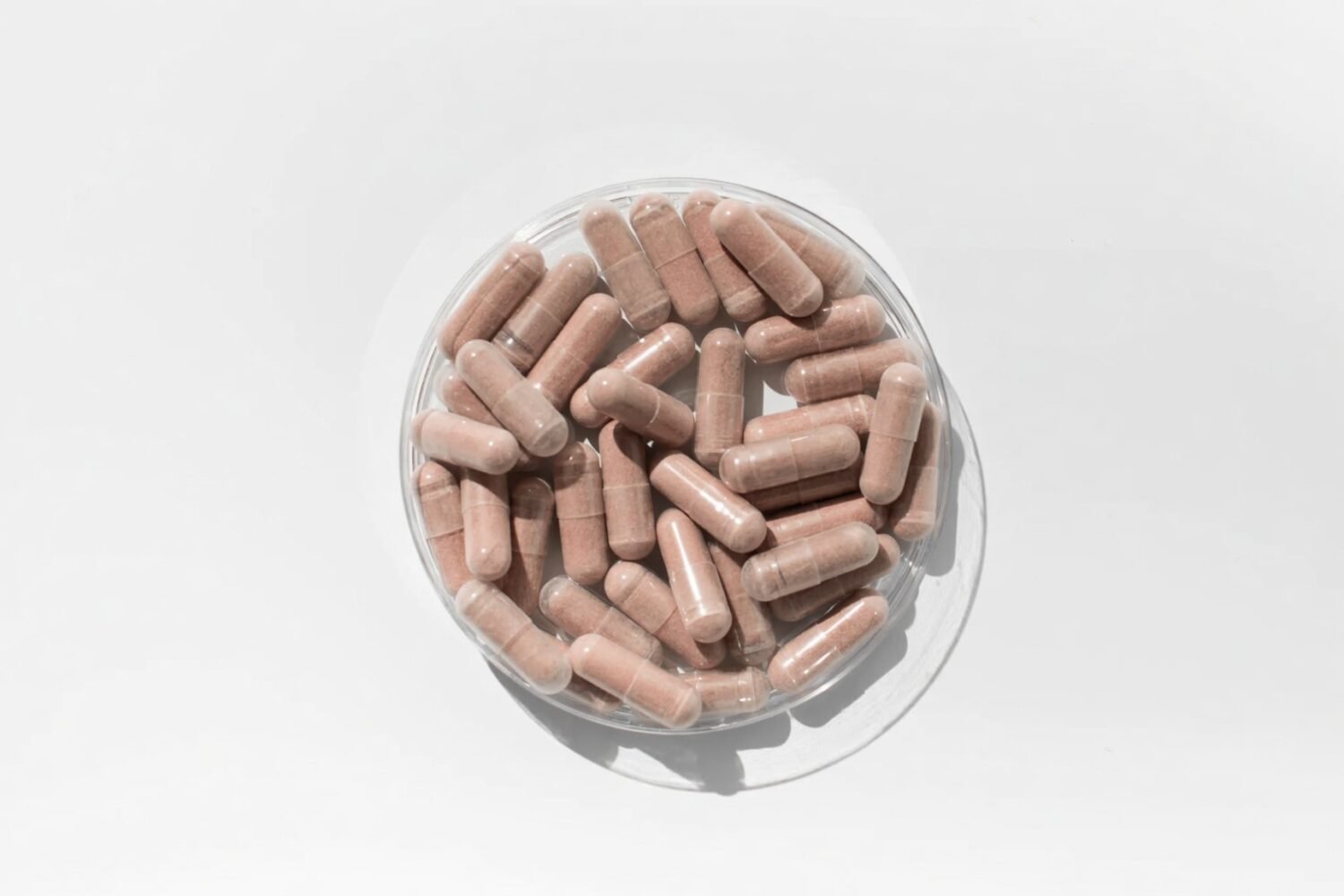 An overhead view of pink-brown supplement capsules piled in a clear glass bowl on a white surface.