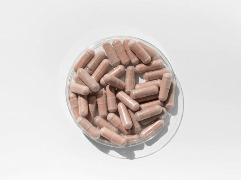 An overhead view of pink-brown supplement capsules piled in a clear glass bowl on a white surface.