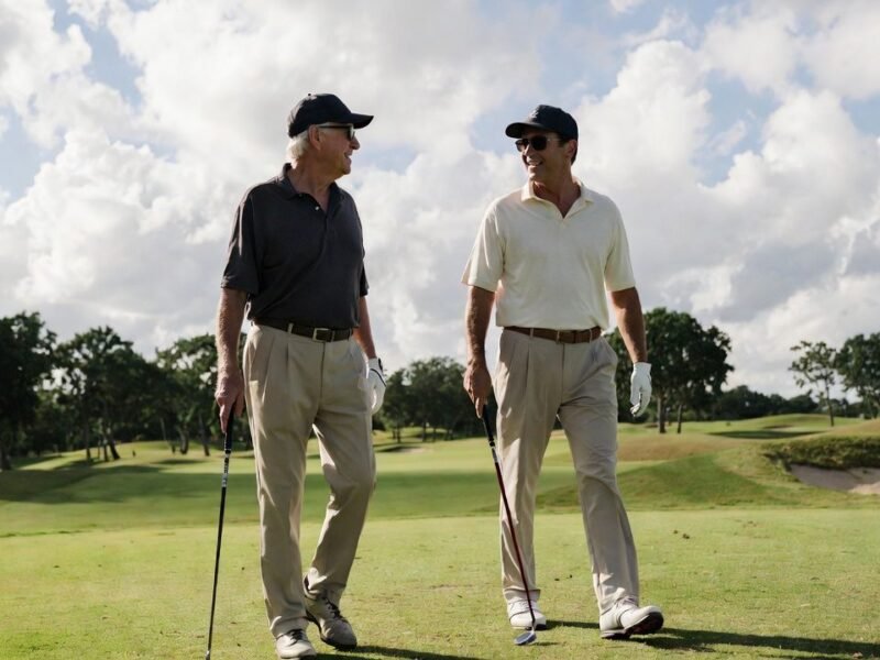 Two men of different ages walk together on a golf fairway under a partly cloudy sky.