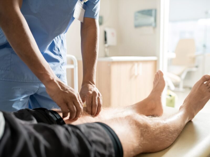 Healthcare worker in scrubs performing a leg examination on a patient lying on a clinical table.